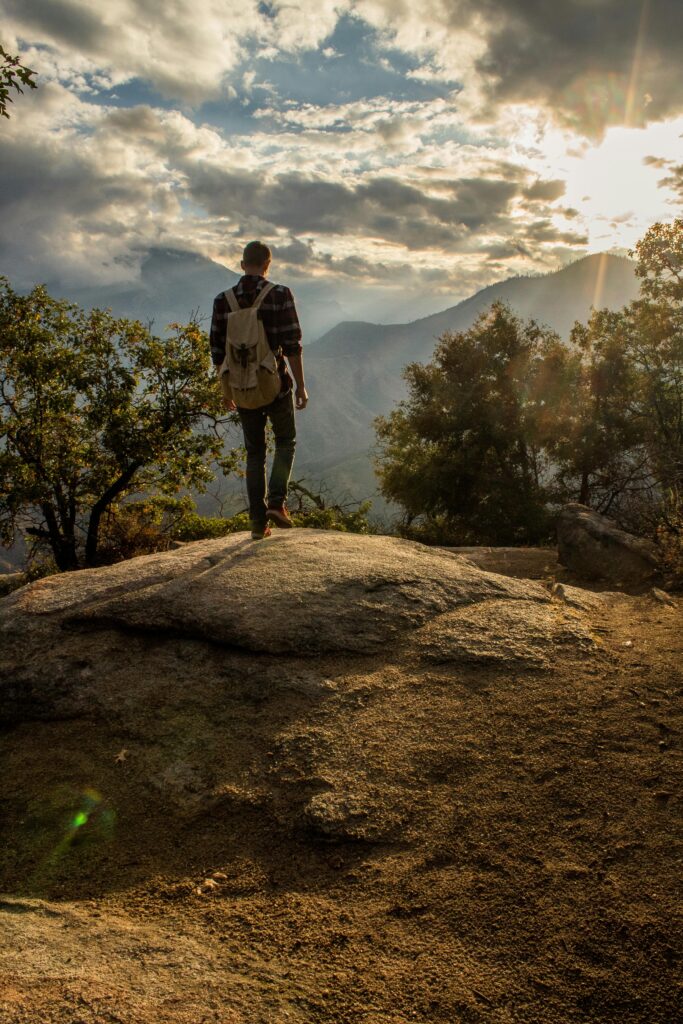 Man hiking on a rocky terrain during sunset, enjoying the scenic view with sun rays breaking through clouds.
